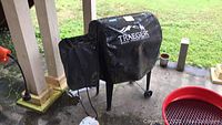 Covered grill under porch showing Traeger logo on cover and attached pellet hopper