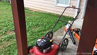 Red Murray push lawn mower shown on grass and concrete, featuring black engine housing, red throttle lever on handlebar, and visible dirt on wheels and mower deck.