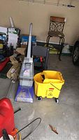 Photo showing Bissell Steam Vac carpet cleaner next to a yellow Rubbermaid mop bucket with wringer in a garage setting.
