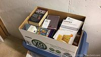 Box of books and office supplies on top of plastic container in basement corner.