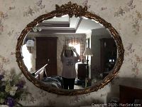 Full view of the large oval shaped mirror with gold decorative trim hung on a floral wallpapered wall, showing part of the room's furniture reflection.