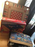 Photo showing stacked items including reversible crokinole board, boxed tabletop billiards game, and wooden train set case on table.