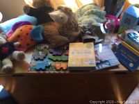 Lot shown on a wooden table includes plush giraffe, wooden animal puzzle, children's books, pink rattle, and other baby items.