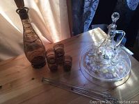 Photo showing both decanter sets with glasses on a wooden table: brown glass decanter with four glasses, clear glass decanter on tray with six glasses, two stirring rods.