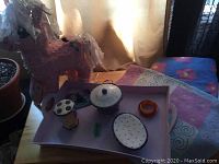 Piñata, purple plastic tray, and three-piece ceramic set on a table with curtains in background