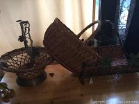 Photo showing three baskets grouped together on wooden surface: oval wicker basket with twisted handle and braided rim, round wicker basket with wrought iron base and handle with leaf details, and rectangular wicker basket with wrought iron handle and grapevine trim.