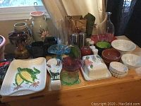 Wide view of collection showing various pottery, glass vases, metal stein, candy dishes, butter dish, salt and pepper shakers, cream and sugar set, platters and bowls on a wooden surface near window.