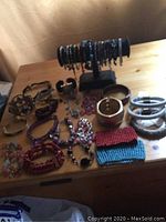Multiple bracelets on a wooden surface including beaded, metal cuff, threaded bead, and leather styles, some displayed on a black bracelet stand.