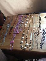 Overview of nine necklaces laid flat on a wooden table, showing a variety of metal and beadwork designs including flower motifs, chunky chains, and purple beads.