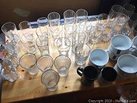 Photo showing various clear drinking glasses and mugs arranged on wooden surface.