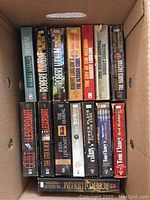 Top view of box containing various paperback books by Robert Ludlum, John Grisham, Tom Clancy, Coonts, and others.