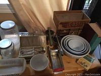 Wide view of the collection on a wooden surface showing jars, silverware, bread pan, mixing bowls, books, and metal canisters.