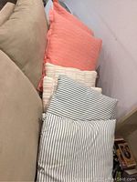 Six cushions stacked together against a beige sofa and wall, showing variety in size and color with peach and striped designs.