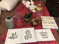 Full lot with three framed notes with text art, faux orchid plant, glass and ceramic vases, glass jug, and watering can on red patterned fabric.