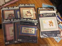 Photo showing multiple Dale Burdett cross stitch kits featuring children, animals, and balloons, unopened and in plastic wrapping
