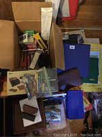 Overview of multiple office supplies including boxed and unboxed desk sets, binders, folders, pencil box, and other items stacked and spread out on a floor