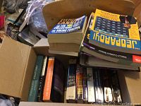 Books stacked in open box and on a chair, featuring a variety of genres including fiction and trivia quiz books.