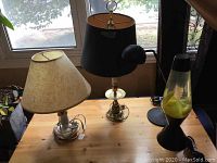 Five assorted lamps on wooden table. From left to right: cream-colored lamp with beige textured shade, brass lamp with black shade and pull switch, yellow lava lamp, black IKEA desk lamp.
