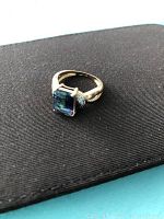 Gold ring with emerald-cut, multicolor mystic topaz centerpiece and small round side accent stones photographed on a black textured surface.