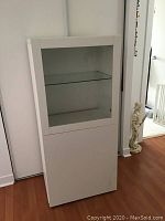 Front view of a tall white storage cabinet with glass door at top and solid door at bottom.