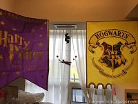 Two banners hanging side-by-side in a room. Left is a purple 'Harry Potter and the Sorcerer's Stone' banner with gold lettering and stars. Right is a yellow banner with the Hogwarts crest, house mascots, and motto.