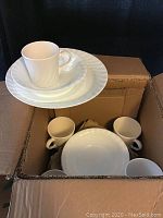 Corelle white dishes stacked in a cardboard box, showing dinners plates, salad plates, bowls and white Corning Ware mugs.