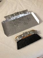 Antique metal crumb collector tray with floral design on front edge and matching brush with wooden handle and floral metal decoration.