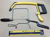 Photo of 3 hack saws and 1 coping saw laid out on beige surface. Includes Stanley 12-inch saw with yellow/black handle and lever, two junior-sized hack saws with black handles, metal coping saw with wooden handle, and a pack of blades.