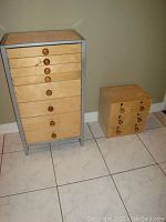 Photo showing the two drawer units side by side on tiled floor against wall. The larger unit has a silver metal frame and 8 drawers with wooden knobs. The smaller unit is plywood with 6 drawers and wooden knobs.