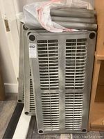 Stacked light gray plastic shelving panels with grid pattern and several plastic cylindrical poles on top, showing components of shelving unit.