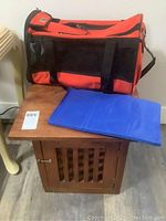 Photo showing wooden pet crate with slatted door, red and black soft pet carrier placed on top, and blue folded pet mat on crate top.