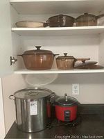 Shelves with multiple amber-tinted glass cookware pieces including covered saucepans and stock pots, stainless steel stock pot and red Salton rice cooker on countertop below.
