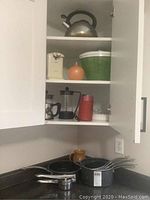 Kitchen shelf with stainless steel kettle on top shelf, white electric can opener, orange ceramic item, green and white salad spinner on middle shelf, Bodum coffee press and grinder on lower shelf, stacked T-Fal pots and pans on counter below