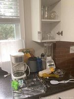 Kitchen counter and cupboard corner showing Black & Decker drip coffee maker with glass carafe, assorted clear glass cups and bowls, hand juicer, blue travel mugs, plastic and metal kitchen utensils including spatula and whisks. Also visible a glass blender pitcher on bottom shelf.