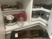 Photo shows open kitchen cabinet with various pots, pans, metal strainer, multiple red-lidded glass and plastic food storage containers, glass mixing bowl, sitting above Salton hotplate on floor.