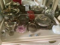 Photo showing a collection of silver plated trays, bowls, salt and pepper shakers, a floral vase with frog wire, and other serve ware pieces on a table.