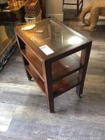 Rectangular wooden side table with inset glass top and two shelves on casters, shown from a front-side angle on wooden flooring.
