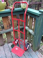 Front view of the red metal hand truck leaning against a green wooden railing outdoors, showing the handle, frame, base plate, and wheels.
