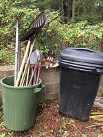 Image showing green plastic garbage can filled with several wooden handled garden tools including rakes, spades, pitchfork, and a large black wheeled garbage can next to it