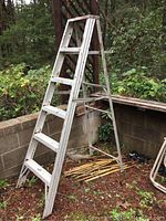 Side view of 6 foot aluminum step ladder showing 5 steps and locking support bar, placed outdoors against a retaining wall.