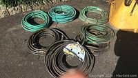 Photo showing seven coiled garden hoses in green and black placed outdoors on pavement, one hose end visibly damaged needing replacement, yellow item partially visible in background.