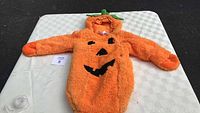 Full front view of orange fluffy infant pumpkin costume with black jack-o'-lantern face and green hood stem/leaf details.