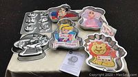 Image showing seven metal cake pans with various children’s birthday themes including princess, cartoon characters, lion, and small animal shapes, arranged on a table.