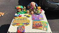 Overview of baby toys on a white table including stuffed puppy, alphabet musical board, piano toy, number puzzle, and books.