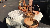 Photo of baskets on table showing large white round wicker basket, tall narrow woven basket, and several other medium and small baskets mostly in natural browns and orange.