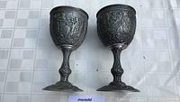 Front view of two pewter goblets showing intricate embossed classical design on bowls and detailed sculpted stems and bases.