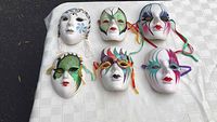Full set of six ceramic Venetian masks laid out on a white cloth with varied colorful designs and attached ribbons