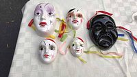 Five ceramic masks arranged on a white textured surface demonstrating size and appearance with colorful ribbons attached.