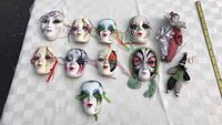 All nine ceramic Venetian face masks and two clown figurines laid out on a white patterned surface with measuring tape alongside for size reference