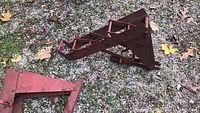Photo of one red metal roof jack lying on gravel with leaf debris, showing triangular base and hooks